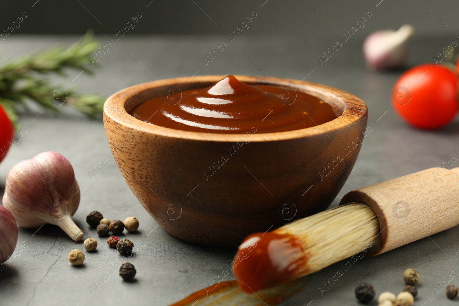 Photo of Tasty barbeque sauce in bowl, brush, garlic and peppercorns on grey table, closeup