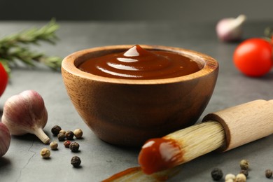 Photo of Tasty barbeque sauce in bowl, brush, garlic and peppercorns on grey table, closeup