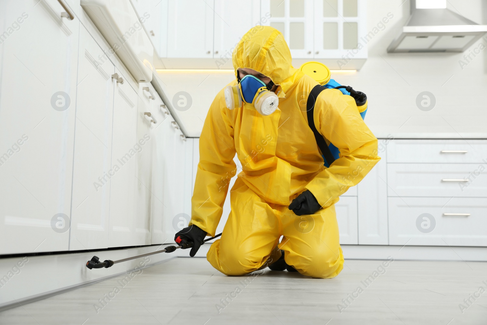 Photo of Pest control worker in protective suit spraying insecticide on furniture indoors