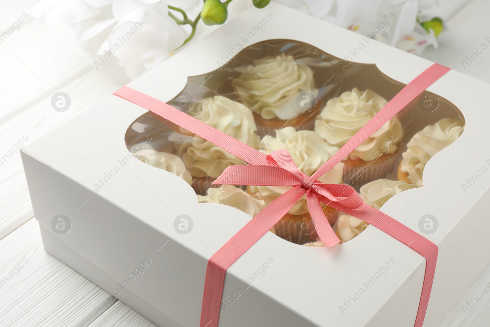 Photo of Tasty cupcakes with vanilla cream in box and orchid flowers on white wooden table, closeup