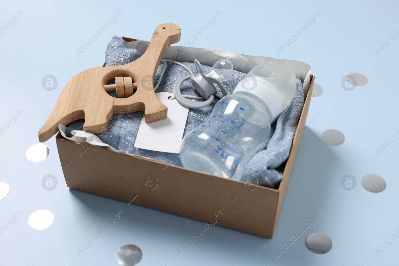Photo of Different baby accessories with blank card in box and confetti on light blue background