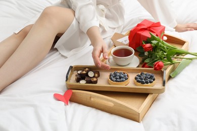 Photo of Tasty breakfast served in bed. Woman with desserts, tea, gift box and flowers at home, closeup