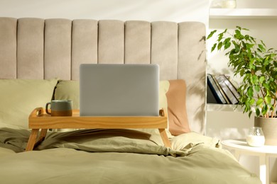 Photo of Wooden tray with modern laptop and cup of aromatic drink on bed indoors