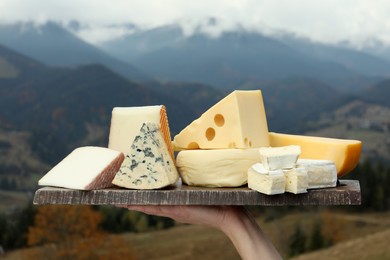 Photo of Woman holding board with different types of delicious cheeses against mountain landscape, closeup