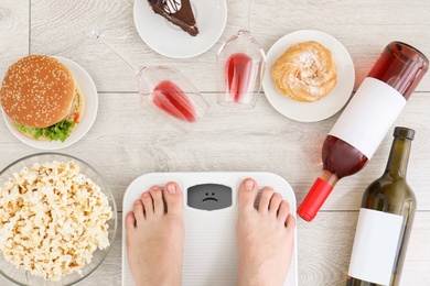 Photo of Woman standing on scales surrounded by different food and alcohol after party indoors, top view