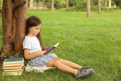 Cute little girl reading book on green grass near tree in park