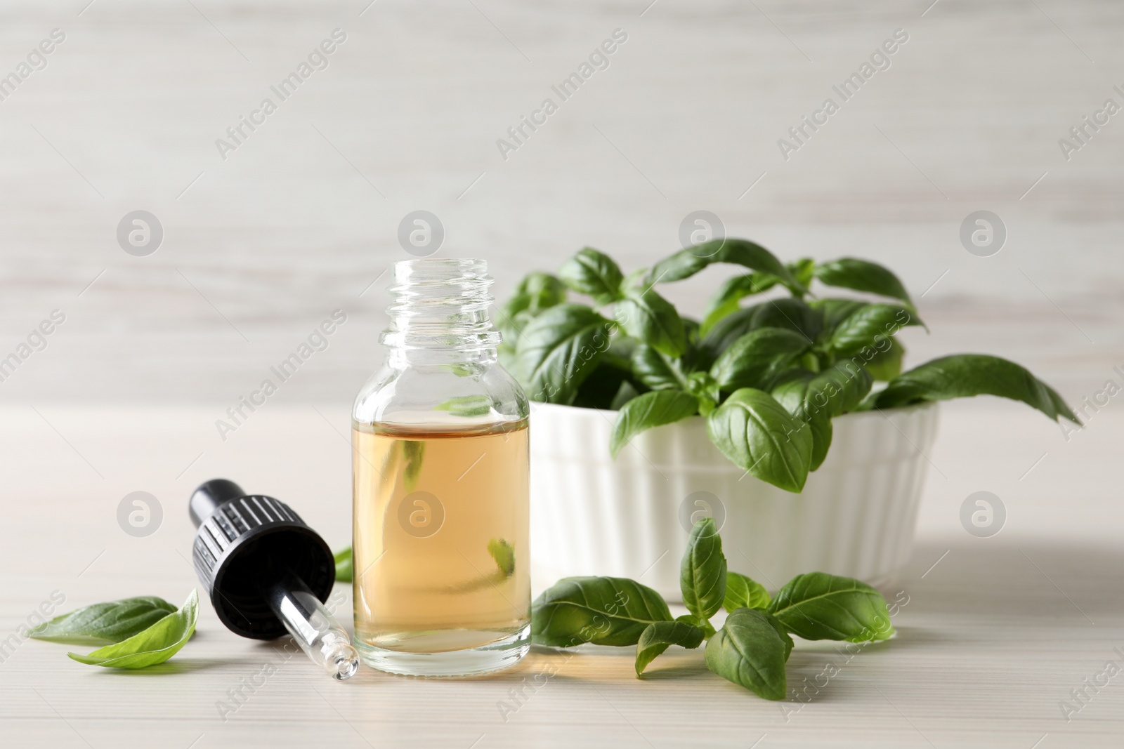 Photo of Glass bottle of basil essential oil near bowl with leaves on white wooden table