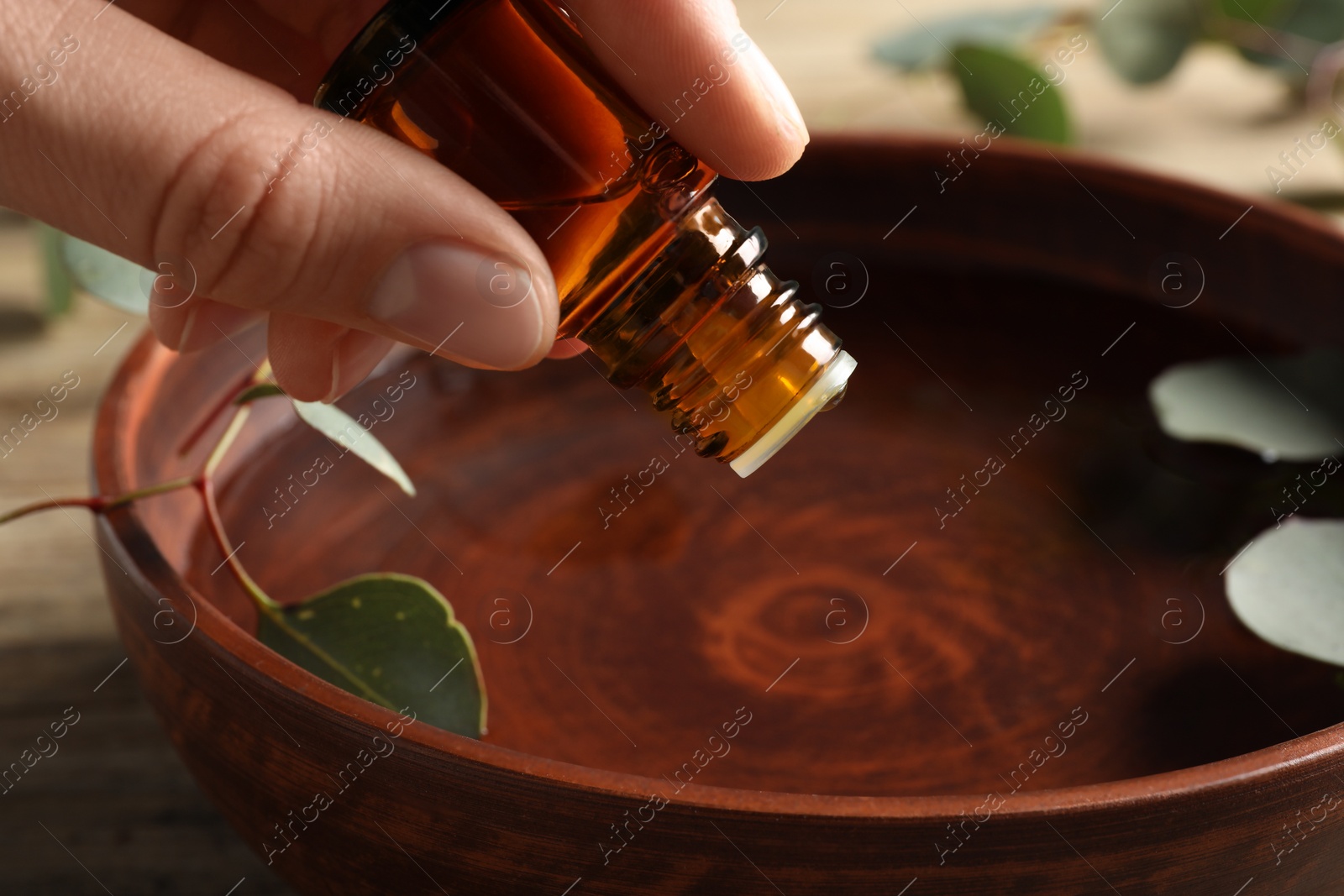 Photo of Woman dripping eucalyptus essential oil from bottle into bowl at wooden table, closeup