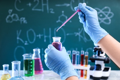 Scientist dripping liquid into bottle against chalkboard, closeup. Chemistry glassware