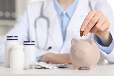 Photo of Doctor putting coin into piggy bank at white table indoors, closeup