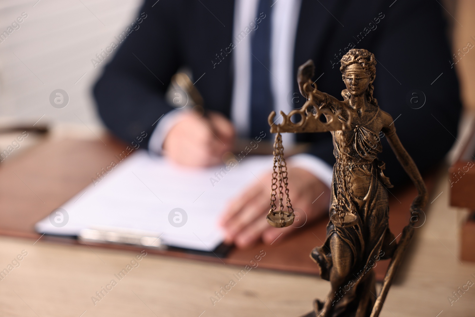 Photo of Notary writing notes at wooden table in office, focus on statue of Lady Justice