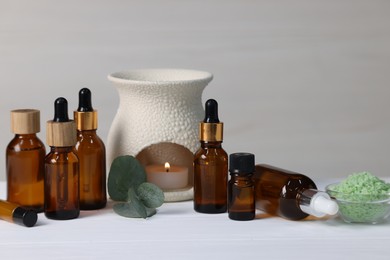 Different aromatherapy products, burning candle and eucalyptus leaves on white wooden table against light background