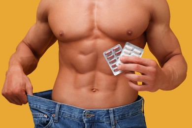 Photo of Athletic man with pills wearing big jeans on orange background, closeup. Weight loss