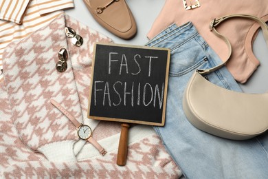 Photo of Small chalkboard with phrase FAST FASHION, stylish clothes and accessories on light background, flat lay