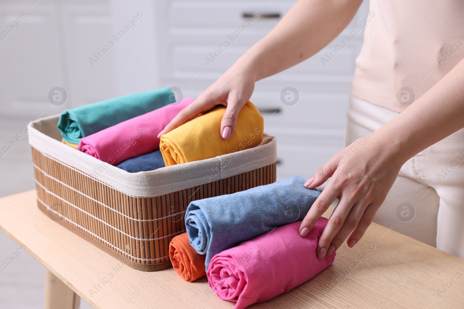 Photo of Woman putting rolled shirt into basket at table in room, closeup. Organizing clothes