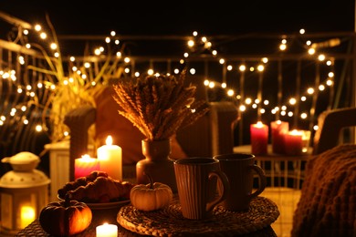 Photo of Rattan furniture, cups, fairy lights, burning candles and other autumn decor on outdoor terrace at night