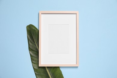 Photo of Empty photo frame and green leaf on light blue background, flat lay. Space for design