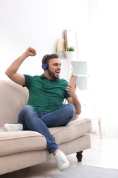 Photo of Young man in headphones with mobile device enjoying music on sofa at home