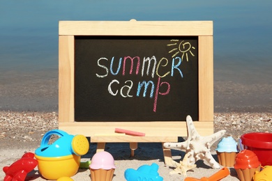 Photo of Small chalkboard with text SUMMER CAMP and beach toys on sand near river