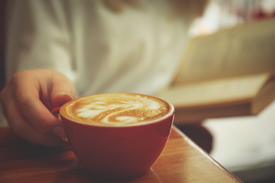 Woman with coffee reading book indoors, focus on cup