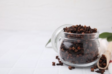 Photo of Aromatic cloves in glass jar and scoop on white tiled table, closeup. Space for text