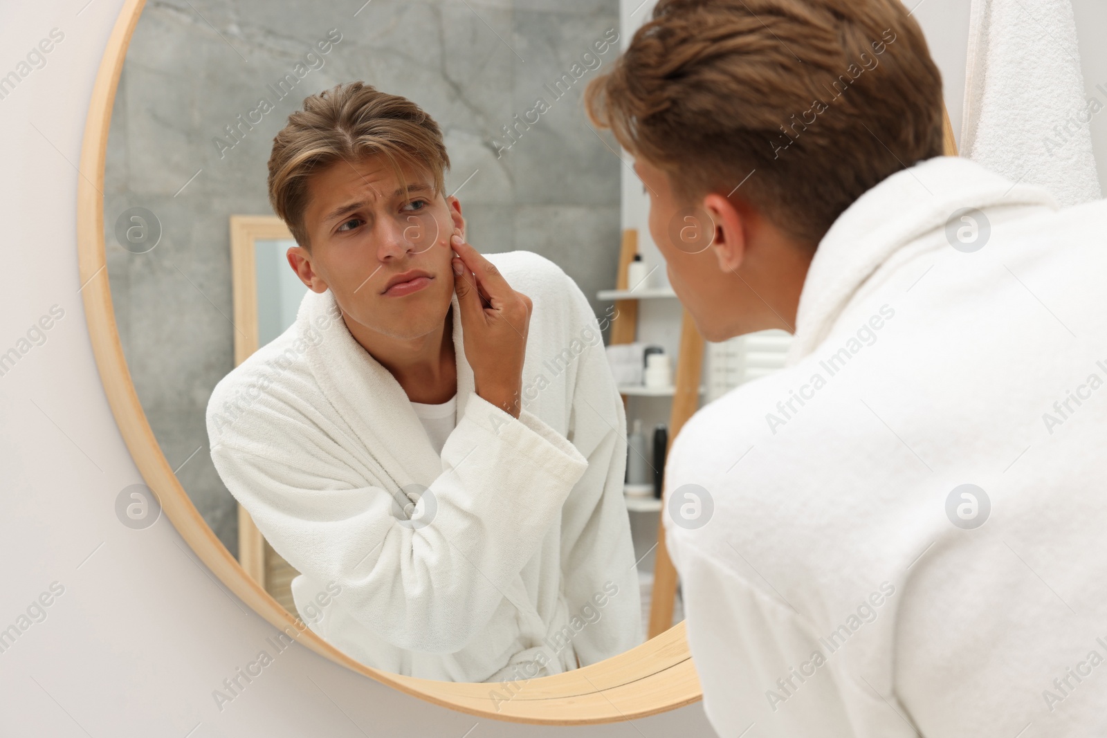 Photo of Upset young man looking at mirror and touching pimple on his face indoors. Acne problem