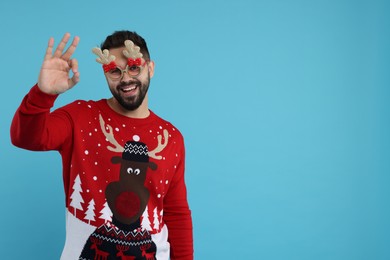 Photo of Happy young man in Christmas sweater and funny glasses showing OK gesture on light blue background. Space for text