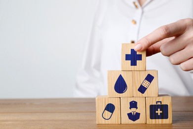 Woman building pyramid of cubes with different icons on wooden table against light background, closeup. Insurance concept