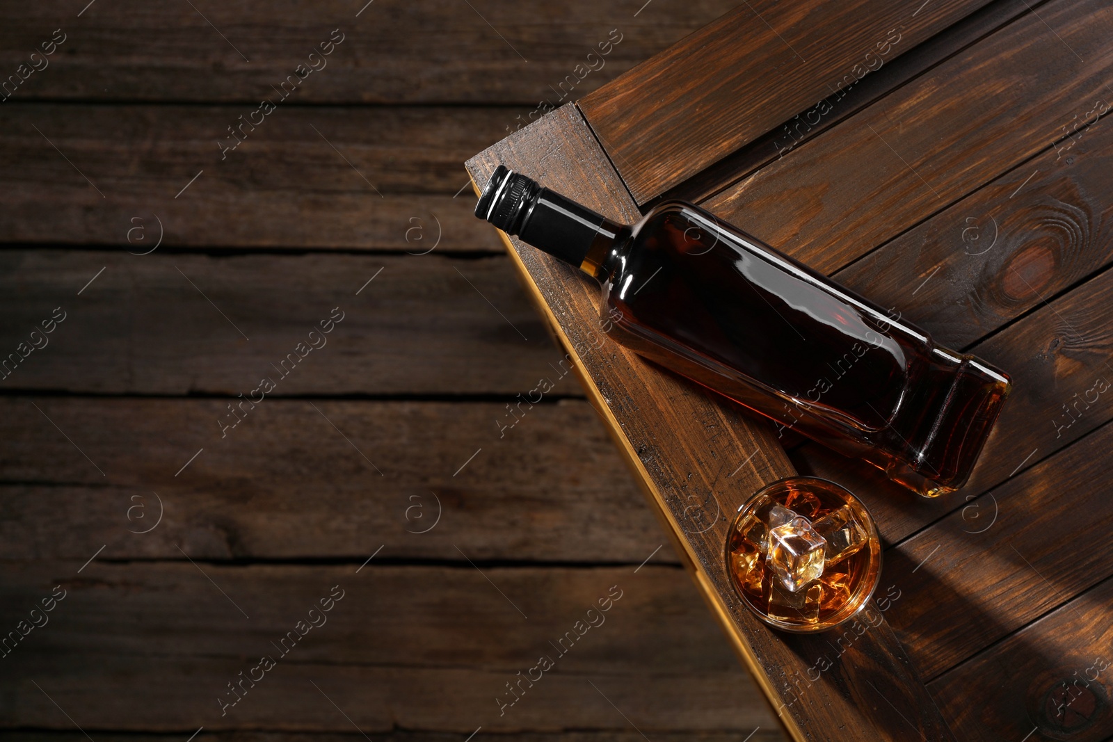 Photo of Whiskey with ice cubes in glass and bottle on wooden crate, top view. Space for text