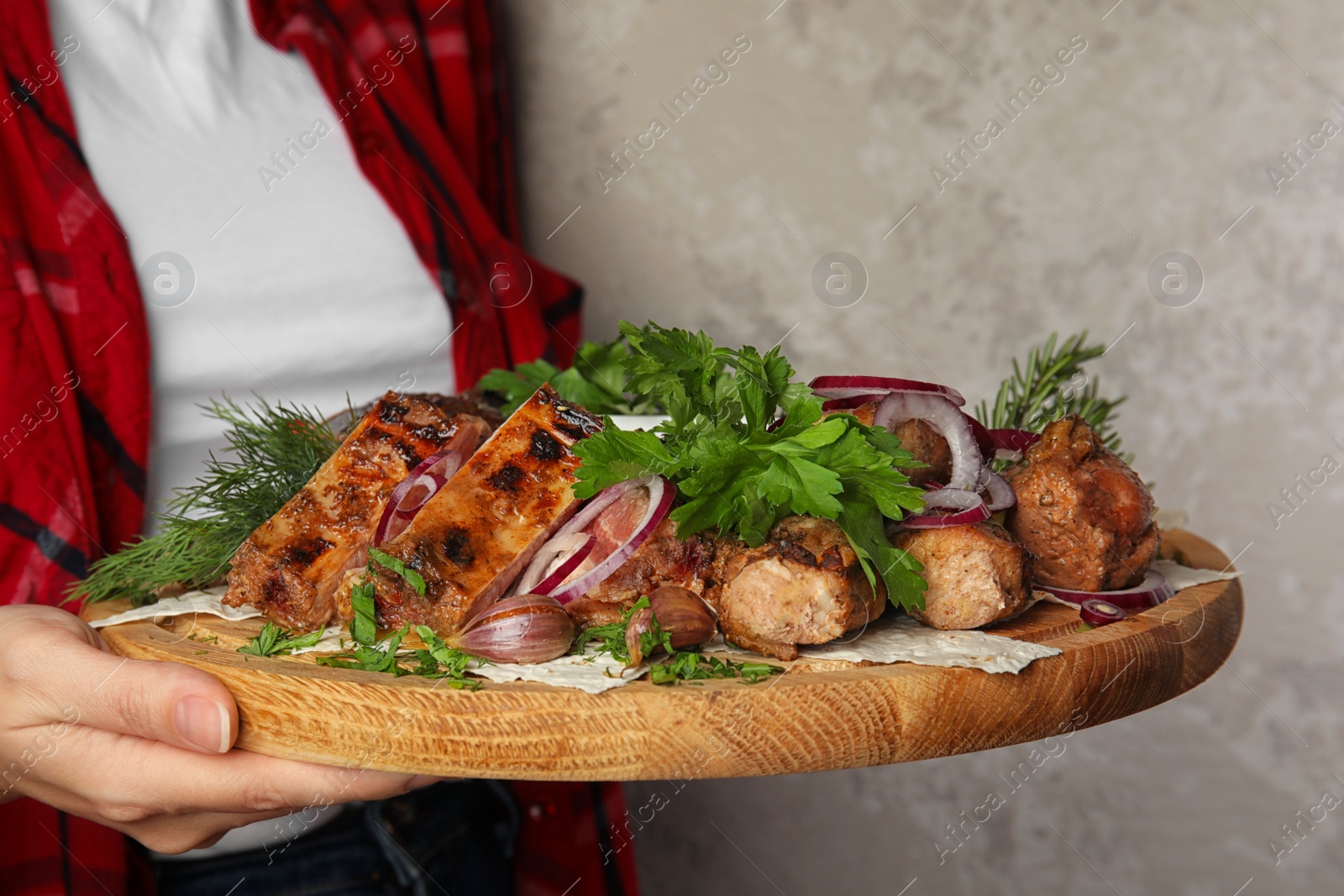 Photo of Woman holding wooden board with delicious roasted meat and herbal on grey background, closeup