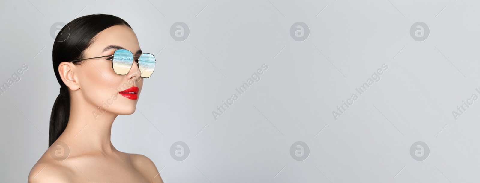 Image of Attractive woman in stylish sunglasses on light grey background, space for text. Sandy beach and wavy sea reflecting in lenses. Banner design