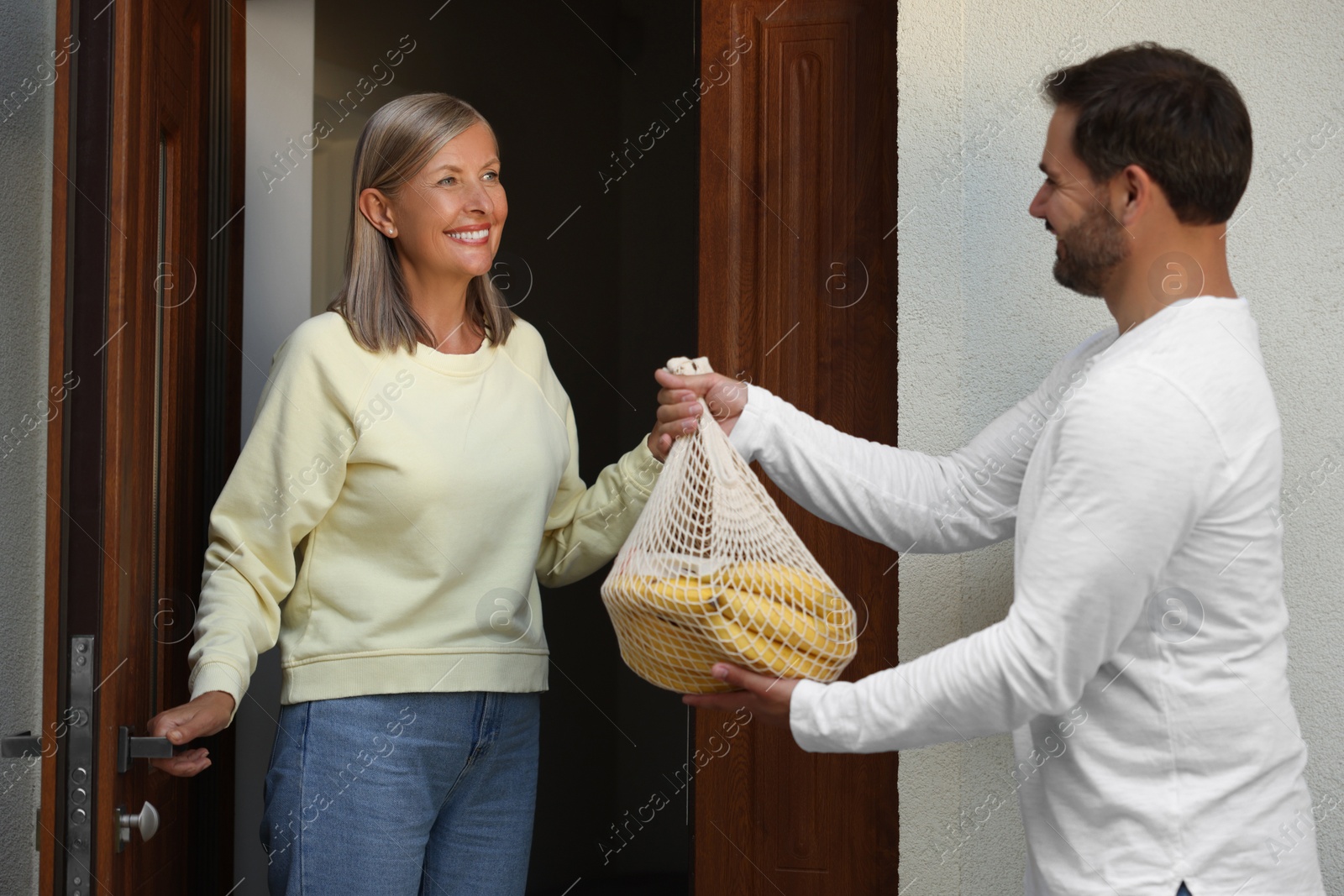 Photo of Helping neighbours. Man with net bag of products visiting senior woman outdoors