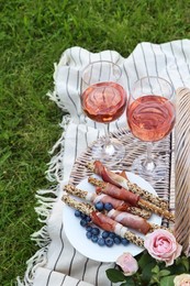 Photo of Glasses of delicious rose wine, food, flowers and basket on picnic blanket outdoors