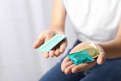 Young woman holding birth control pills and condoms on light background, closeup. Safe sex concept