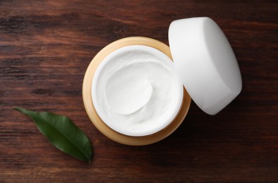 Jar of face cream and green leaf on wooden table, top view