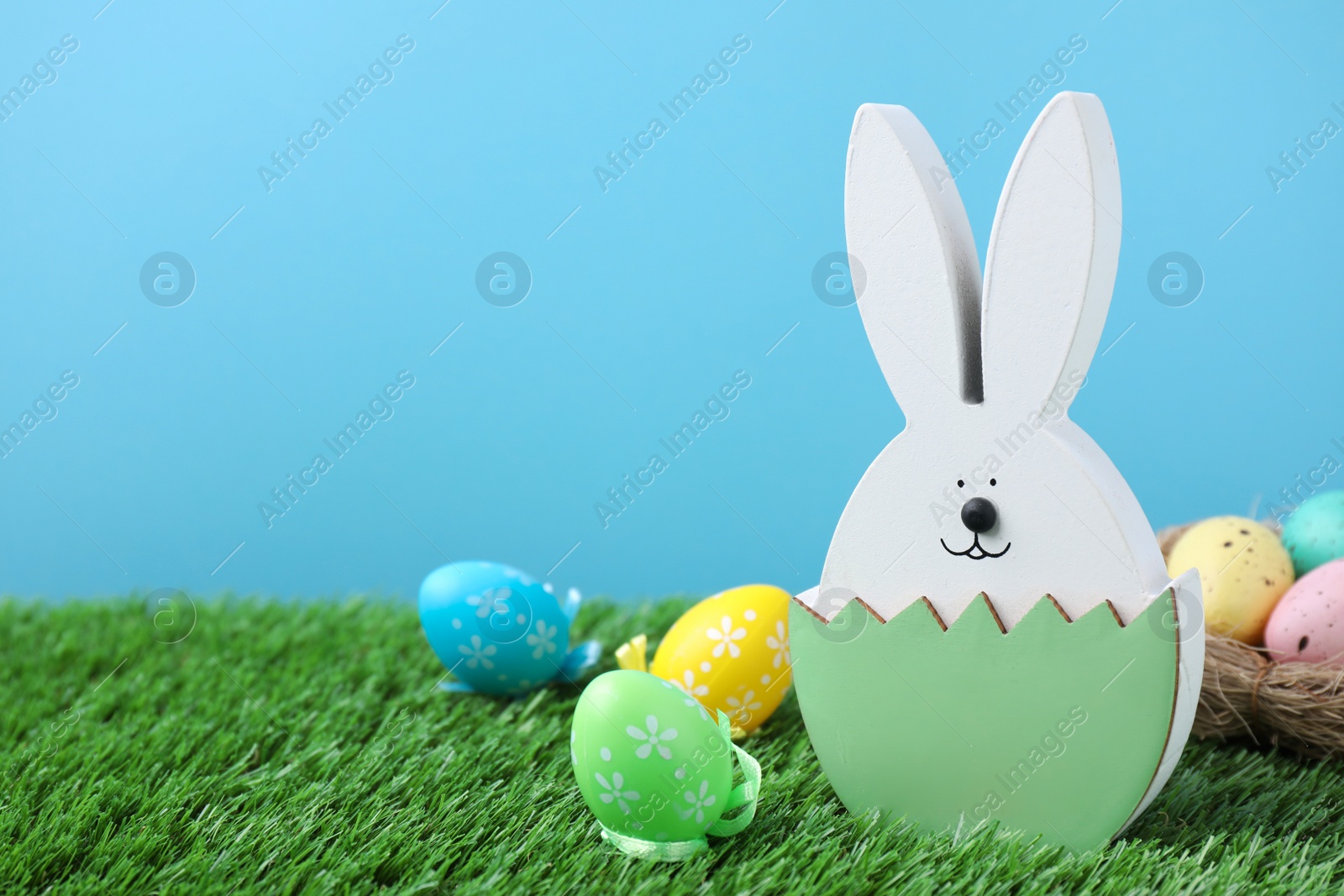 Photo of Easter bunny figure and dyed eggs on green grass against light blue background. Space for text