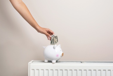 Photo of Woman putting money into piggy bank on heating radiator against light background