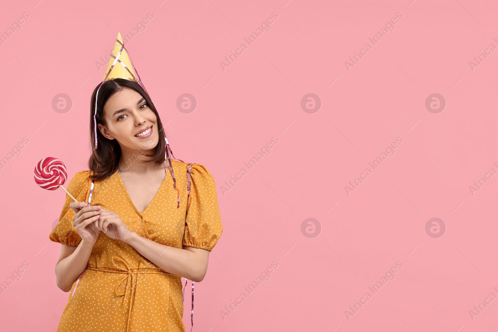 Photo of Happy young woman in party hat with candy on pink background