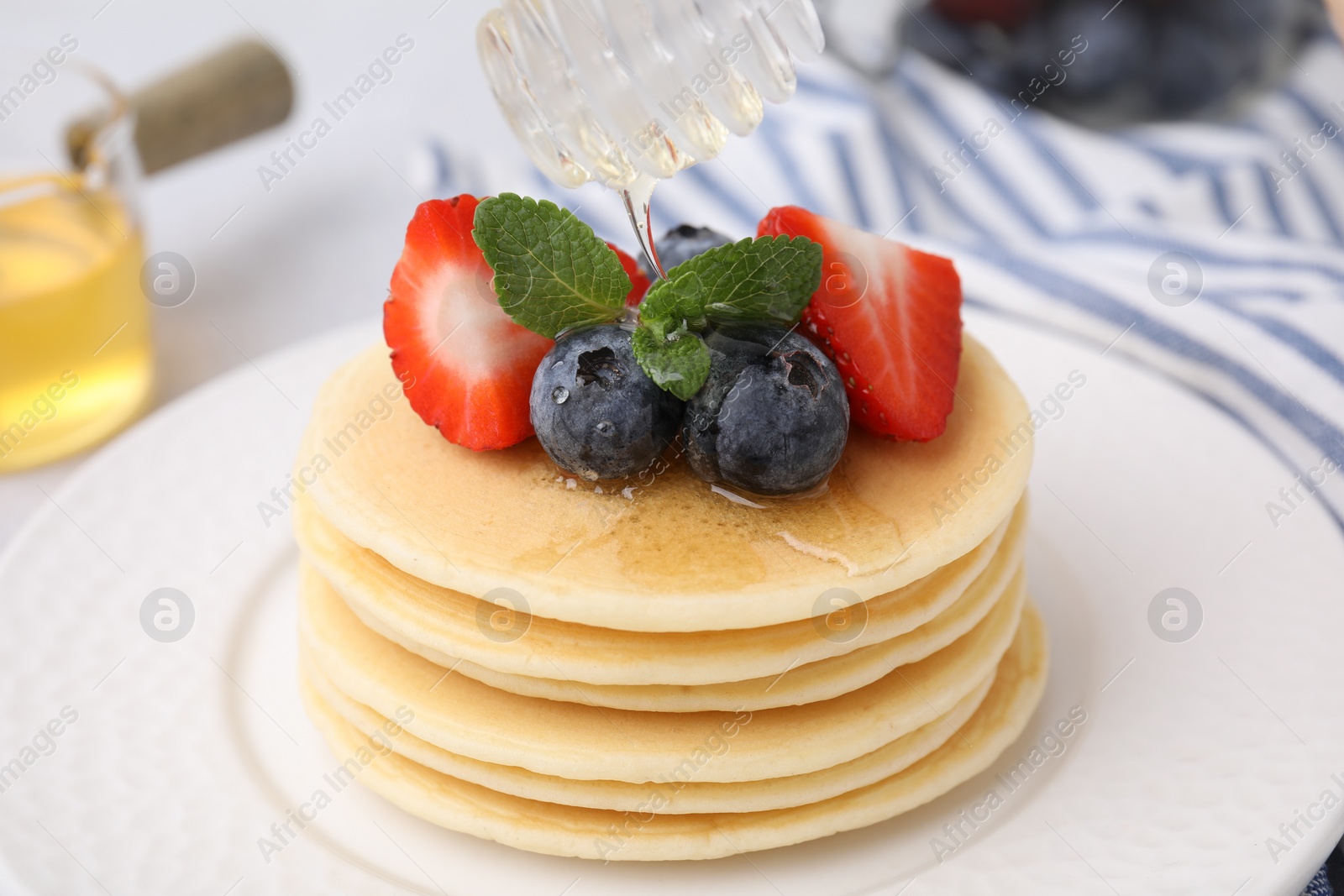 Photo of Pouring honey from dipper onto delicious pancakes with strawberries, blueberries and mint at table, closeup