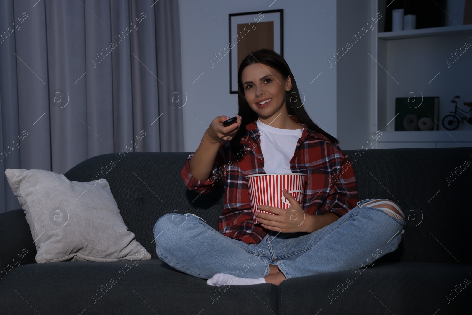 Photo of Happy woman holding popcorn bucket and changing TV channels with remote control at home in evening