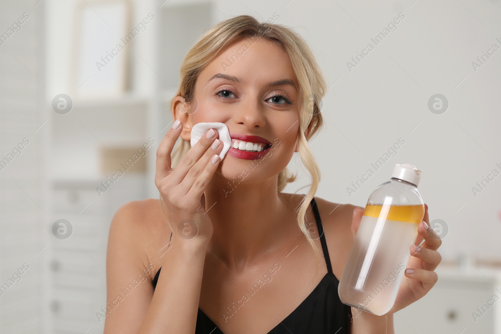 Photo of Smiling woman removing makeup with cotton pad and holding bottle indoors