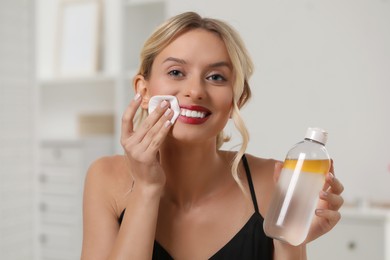 Smiling woman removing makeup with cotton pad and holding bottle indoors