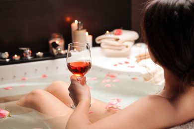 Photo of Woman holding glass of wine while taking bath with rose petals, back view. Romantic atmosphere