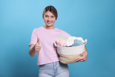 Photo of Happy woman with basket full of laundry showing thumb up on light blue background
