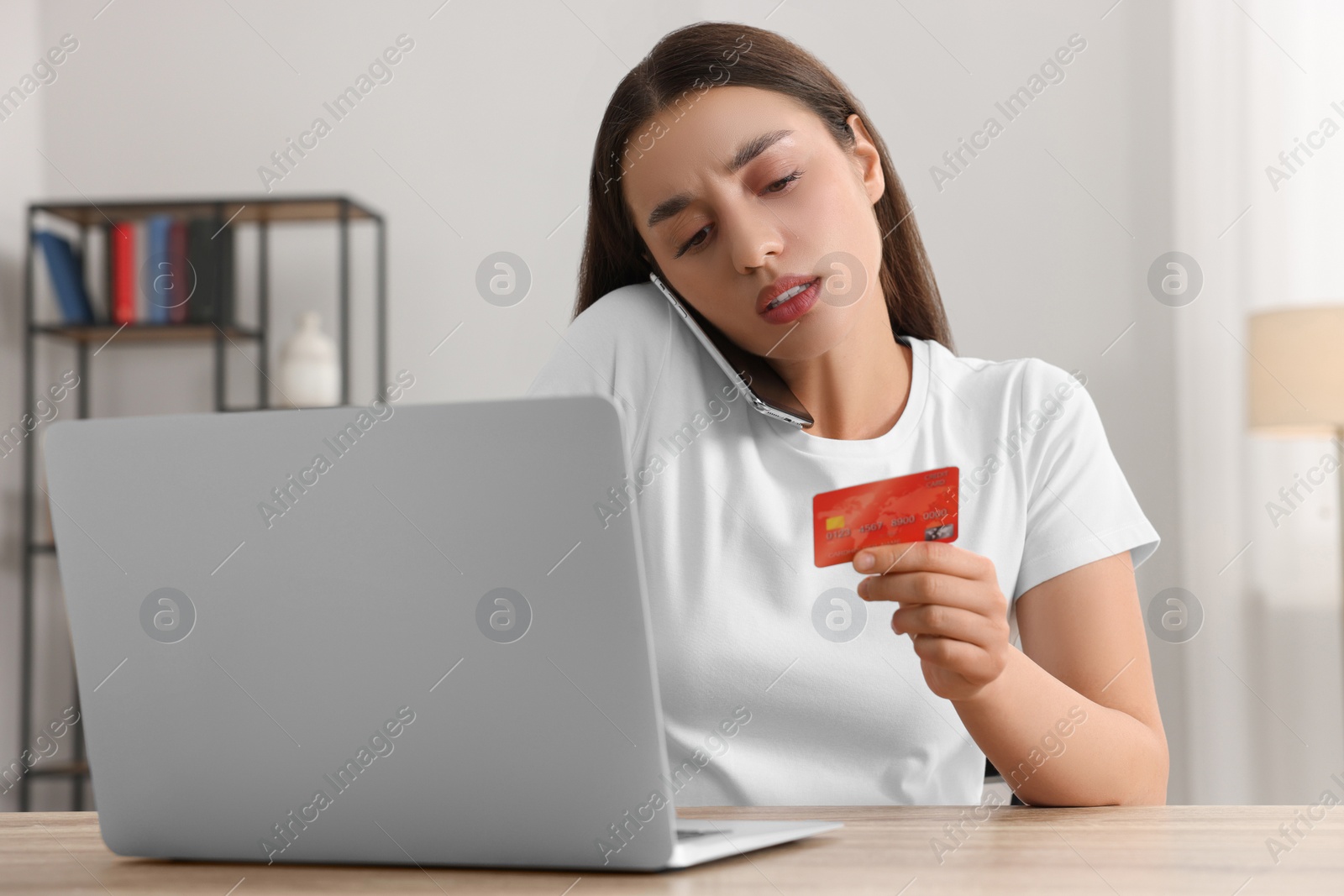 Photo of Confused woman with credit card talking on phone and using laptop at table indoors. Be careful - fraud