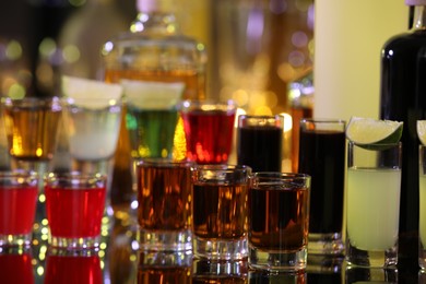 Photo of Different shooters in shot glasses and bottles on mirror surface against blurred background. Alcohol drink