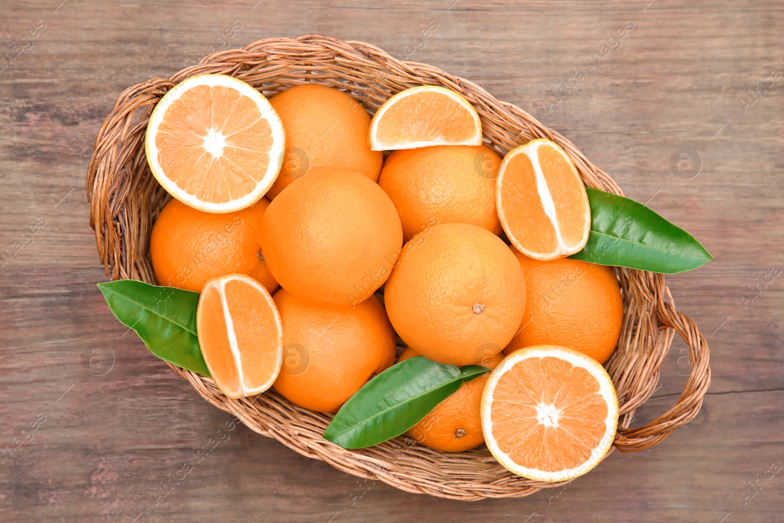 Photo of Wicker basket with ripe juicy oranges and green leaves on wooden table, top view