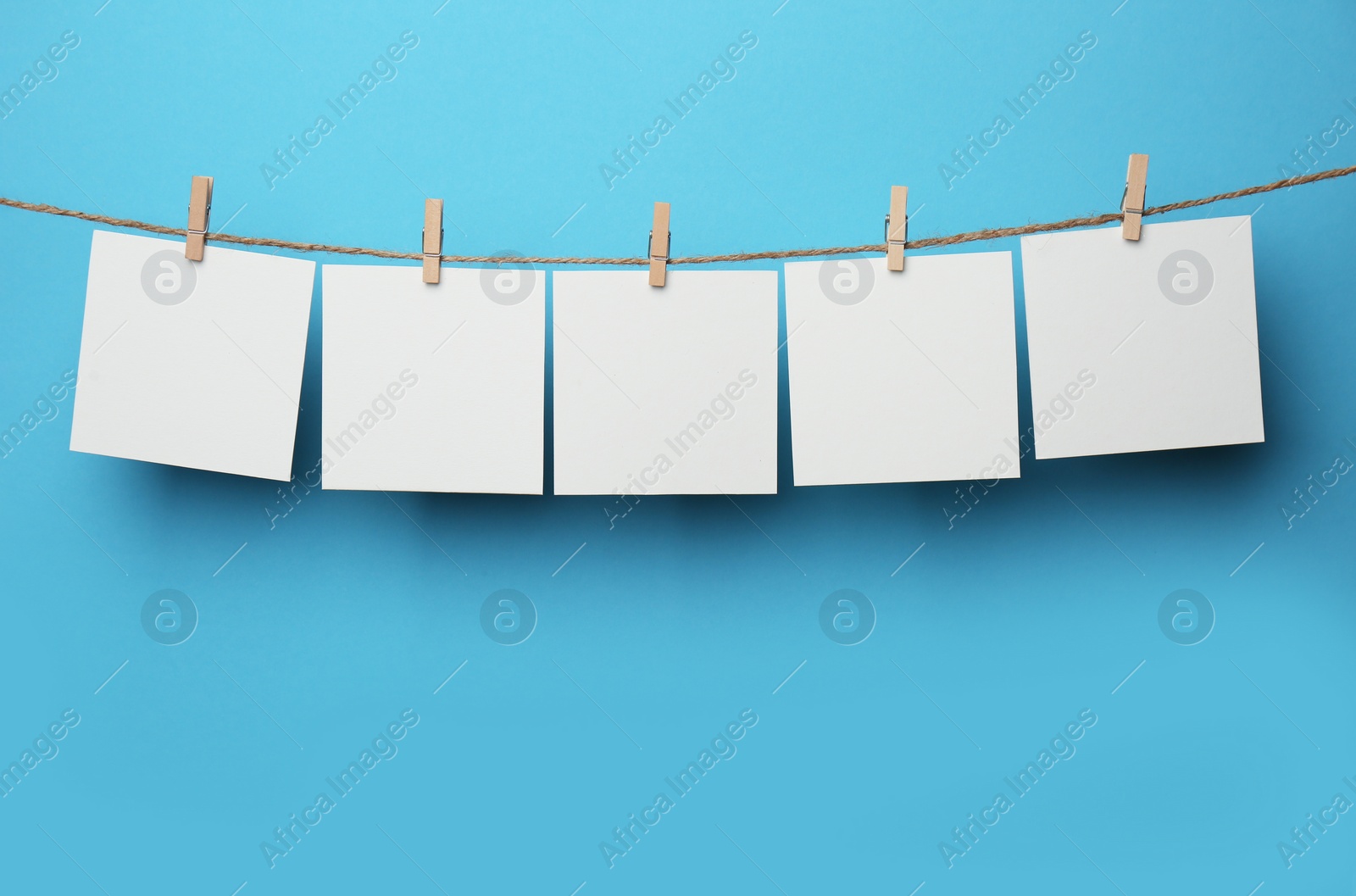 Photo of Wooden clothespins with blank notepapers on twine against light blue background. Space for text