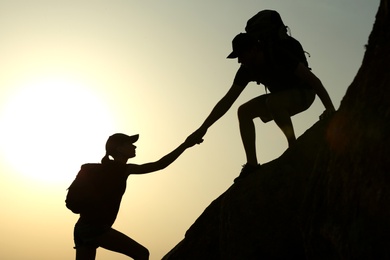 Photo of Silhouettes of man and woman helping each other to climb on hill against sunset