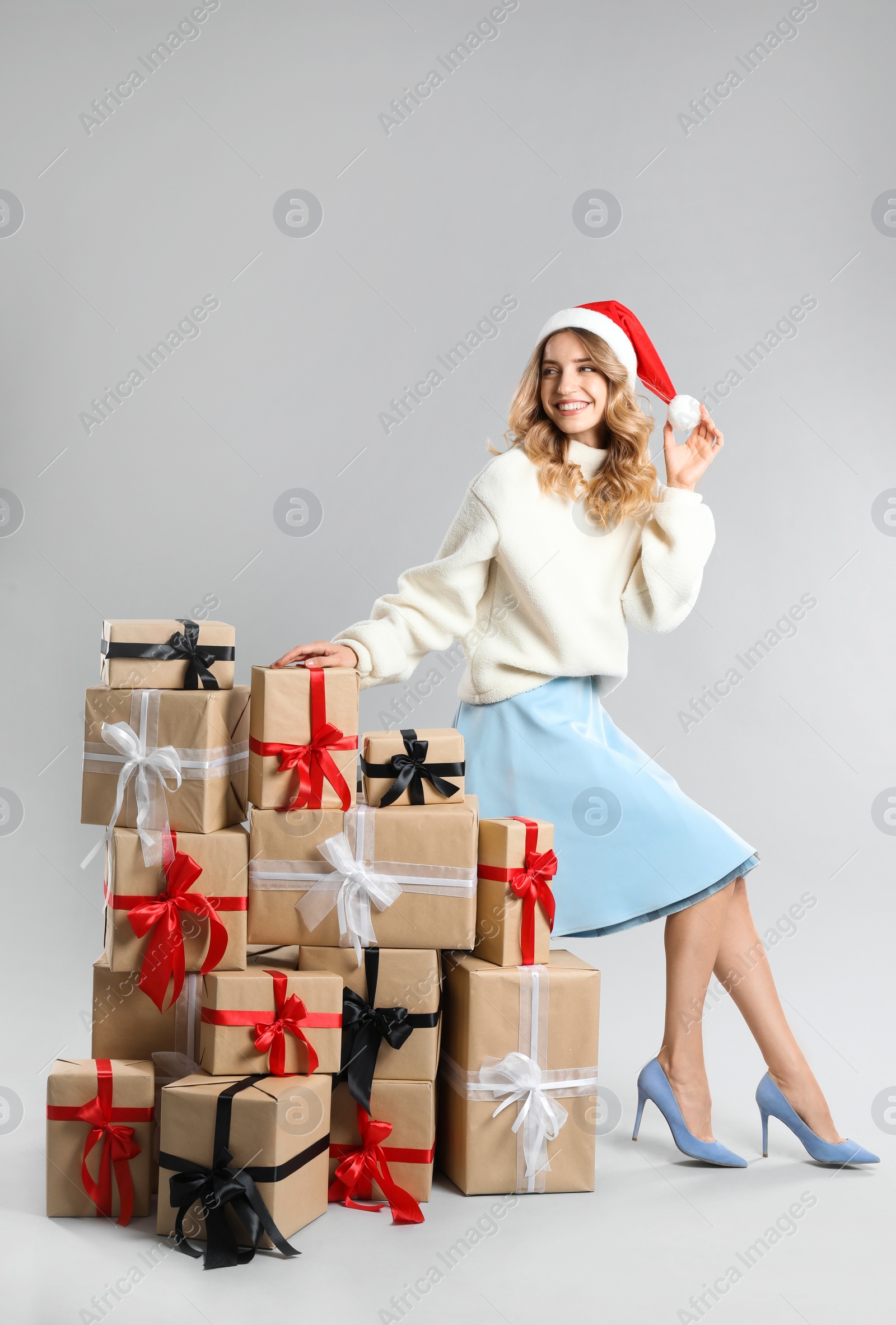 Photo of Beautiful young woman in Santa hat with pile of Christmas presents on light grey background